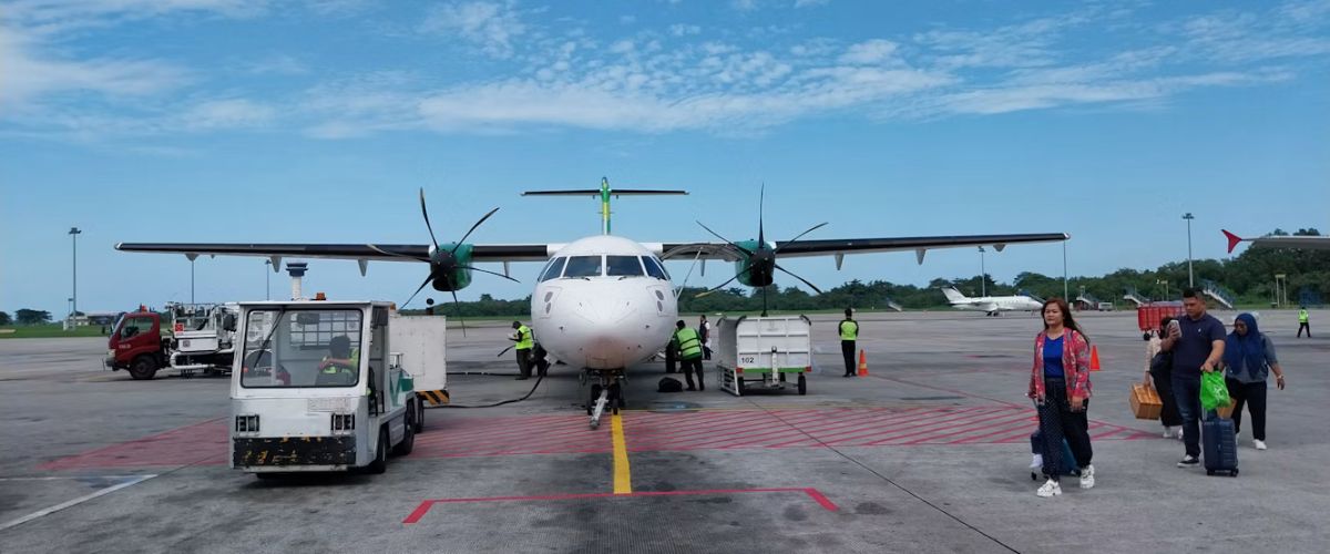 Passengers walking across the tarmac after disembarking from a twin-propeller turboprop plane parked at a regional airport under a clear blue sky.